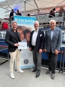 Drei Männer lächeln in die Kamera, im Hintergrund ist ein Schiff mit Personen darauf zu erkennen und direkt hinter der Gruppe ein "Kiwanis International"-Rollup. Der Mann links hält einen kleinen symbolischen Spendenscheck in die Kamera. Es handelt sich um Kai Gosslar und Peter Keller mit Dr. Arthur Friedrich vom Verwaltungsrat der Pestalozzi-Stiftung Hamburg.