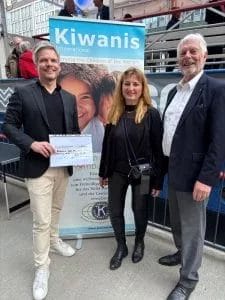 Eine Frau und zwei Männer lächeln in die Kamera, im Hintergrund ist ein Schiff mit Personen zu erkennen und direkt hinter der Gruppe ein "Kiwanis International"-Rollup. Der Mann links hält einen kleinen symbolischen Spendenscheck in die Kamera. Es handelt sich um Kai Gosslar von Lecker³ und der Pestalozzi-Stiftung Hamburg mit Felicitas von der Burg und Peter Keller von den Kiwanis Hamburg.